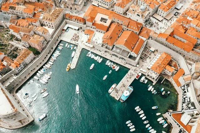 Aerial view of boats docked in a historic harbor surrounded by orange-roofed coastal buildings.